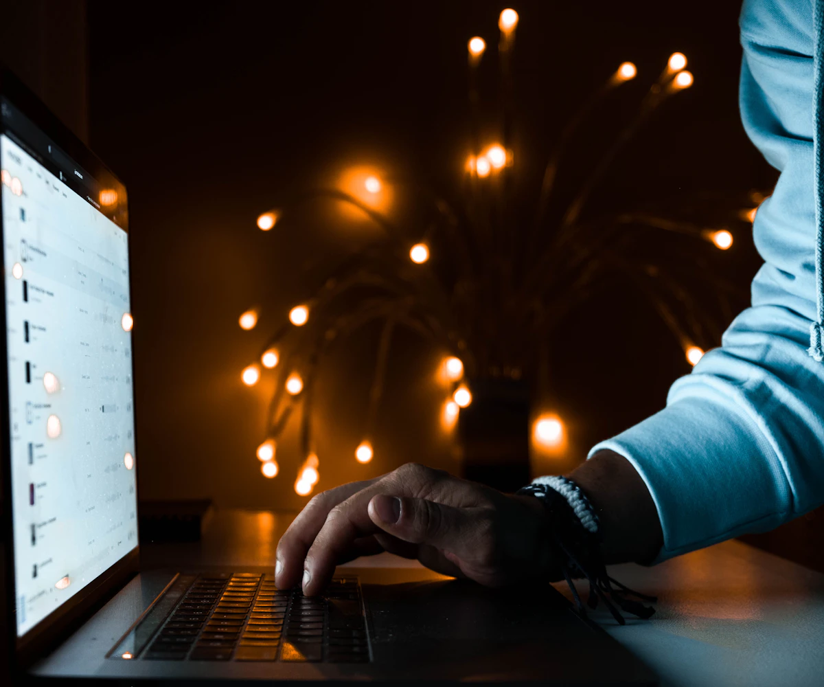 Hand typing on a laptop keyboard at night with warm lights glowing in the background